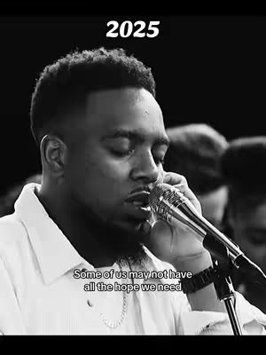 A black and white image of a Black man singing into a microphone, with text overlays. The man has a short fade haircut, wears a button-down shirt, and a chain necklace. The background is blurred, suggesting a performance or studio setting. Text at the top reads '2025', and a quote at the bottom says, 'Some of us may not have all the hope we need'. The image is well-lit and emotionally charged.