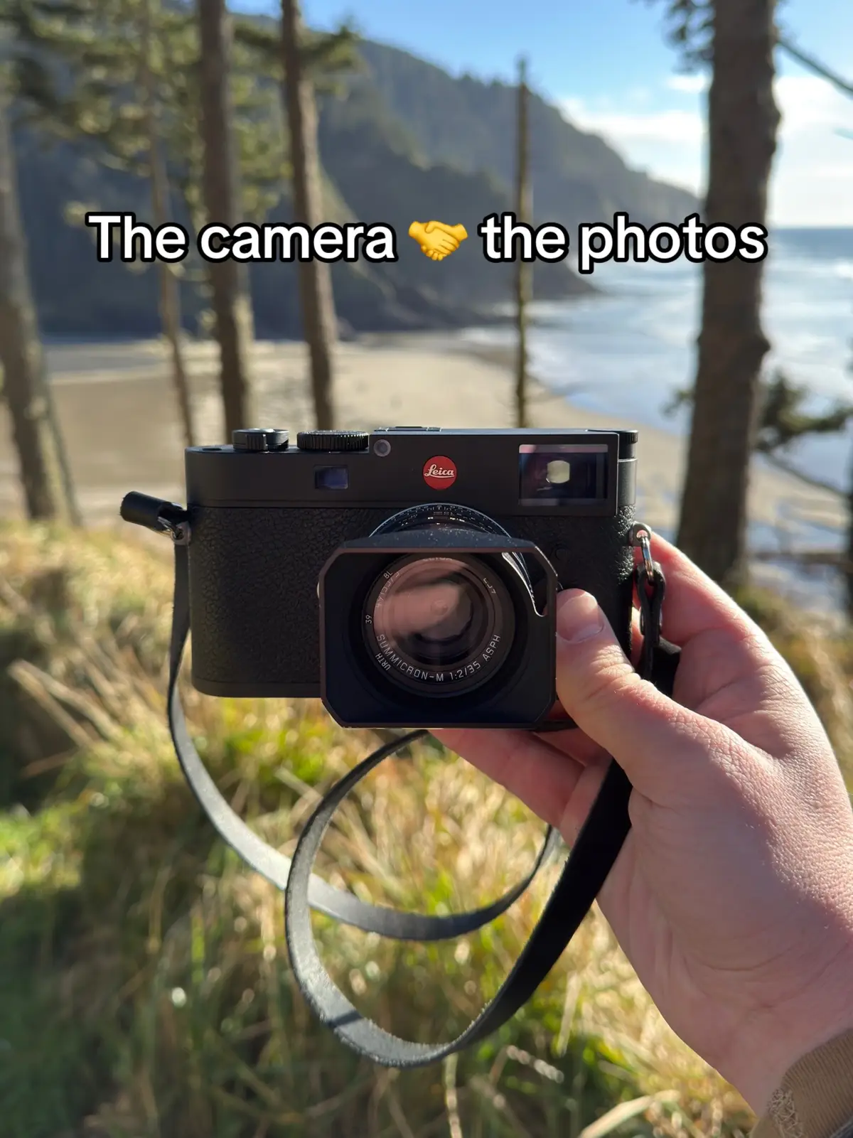A person holds a black Leica camera in front of a scenic background featuring a beach, ocean, and trees. The camera lens is prominent, and text overlays the image.