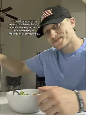 A young man is seated at a table, holding a bowl of food. He's wearing a black baseball cap with an American flag on it and a light blue t-shirt. The background is blurred, with a ceiling fan visible. Text overlayed on the image expresses disappointment and a lack of motivation.
