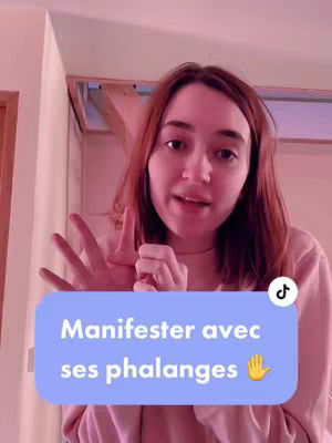 A young woman with light skin and reddish-brown hair is centered in the frame, speaking directly to the camera while gesturing with her hands. A blue bubble highlights the text 'Manifestrer avec ses phalanges' and a hand emoji. The background is a soft pink.