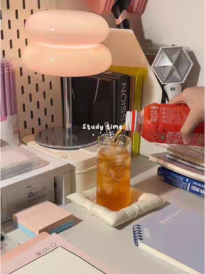 A well-lit photo of a desk setup with studying supplies. In the foreground, a glass of amber liquid sits atop a small white pillow, with ice cubes visible. A red bottle is pouring liquid into the glass . In the background, a rounded, pink-toned lamp dominates. Besides, there are also a hexagonal light, notebooks, and sticky notes are visible. The overall aesthetic is soft and inviting.