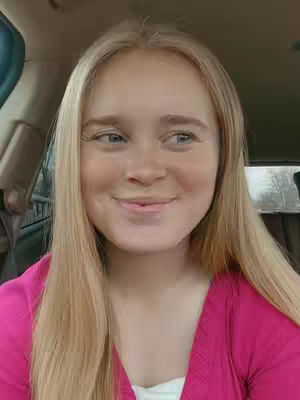 A woman with long blonde hair is sitting in a car, looking at the camera with a slight smile. She is wearing a pink shirt.