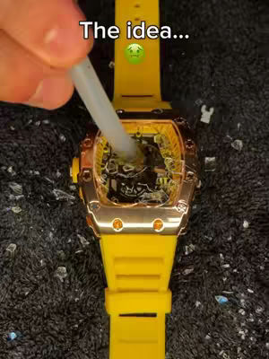 The image showcases a close-up shot of a luxury watch. A hand, visible in the upper left, is inserting a white tube into the watch's mechanism. The watch features a yellow strap and a transparent face, revealing the inner workings. The background is a dark, textured surface, possibly fabric or a similar material, with shards of glass-like material scattered around the watch.