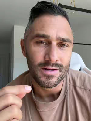 A medium close-up shot of a man looking directly at the camera with a slight smile. He has short dark hair with a subtle fade on the sides and a short beard. He is wearing a brown t-shirt. The background is blurred, suggesting an indoor setting.