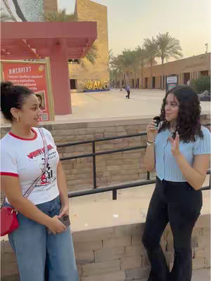 Two young women are standing in what appears to be a public square or university campus. One is holding a microphone and gesturing, while the other is facing her, looking at her and smiling. The backdrop includes buildings with arches and a sign.