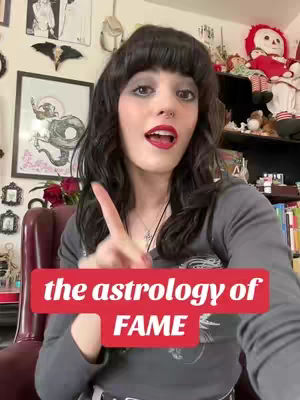 A woman with dark hair and red lipstick is pointing upwards with her right index finger. She is wearing a dark gray shirt and a silver necklace. A red banner across the lower portion of the frame reads 'the astrology of FAME.' The background includes framed artwork, decorative items, and shelves with various objects.
