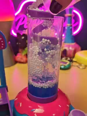The image shows a close-up of a clear, cylindrical container filled with a spiral structure. White, ball-like objects are cascading down the spiral. The container sits atop what appears to be a pink base, possibly a donut shape, with a blue bottom. The background includes blurred, colorful elements, including a neon sign.