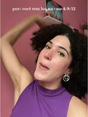 A woman with dark, curly hair and wearing a purple top is angled toward the camera, her arm is raised, and the background is a pink wall with an image and shelf.