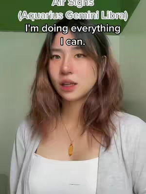 A young woman with shoulder-length brown hair is centered in the frame, looking directly at the camera with a neutral expression. Above her, the text overlay indicates the video's subject matter: Air Signs (Aquarius Gemini Libra) and a phrase, "I'm doing everything I can." The background appears to be a simple light green surface. She wears a white top with a silver necklace with a pendant.
