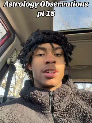 A selfie-style shot inside a car. The main subject, an African-American male with short locs, fills much of the frame. He wears a dark, textured hoodie and looks directly at the camera with a slight smile. The background includes the car's interior, parts of the windshield, and a glimpse of sunlight and trees. The image has well-lit and balanced, appearing natural with a slightly warm tone.