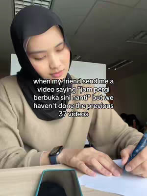 A woman wearing a black hijab and a beige sweater is seated and writing on a piece of paper with a pen. In the background, there are office furnishings.