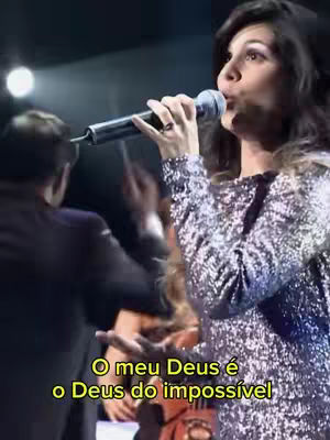 A woman with dark hair holding a microphone and singing on stage with a crowd in the background. Text overlayed on the image reads, "O meu Deus é o Deus do impossível."