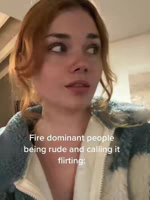 A woman with reddish-brown hair is centered in the frame, looking off to the side with a slightly inquisitive expression. Text overlayed near the bottom reads 'Fire dominant people being rude and calling it flirting:'. She wears a light blue, textured jacket.