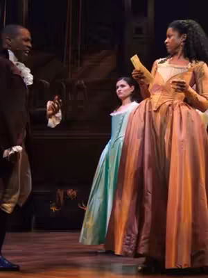A stage scene from the musical Hamilton, featuring three actors in period costumes. One person is speaking, holding a paper. The background is a dimly lit theater setting.