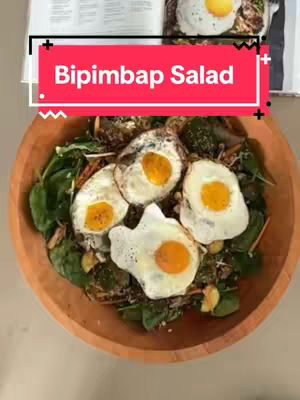 A wooden bowl filled with a salad consisting of spinach, fried eggs, and other ingredients. The text overlay reads 'Bipimbap Salad' in a red box.