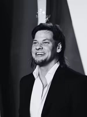 A grayscale image of a man smiling. He has dark hair, a small beard, and is wearing a suit jacket over a white shirt. Above his head are two stars and the words "BE SELF" written in an artistic font. The background is blurred and appears to be an indoor setting.