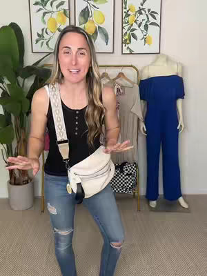 A woman stands in a well-lit room, gesturing with both hands. She wears blue jeans with distressed details and a black top. A white crossbody bag with a patterned strap is across her chest. Behind her, a clothing rail displays garments, and two framed lemon-themed art pieces are on the wall.