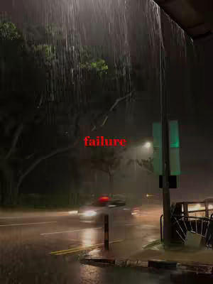 A nighttime street scene during a heavy rainstorm. Rain streaks down from the top of the frame, with a street lit by streetlights, headlights from passing cars. A sign, a traffic light, and a bus stop are on the right side, partially obscured by the rain and darkness. The image has a moody, atmospheric feel with dark tones. In the foreground, pavement wet from the rain.