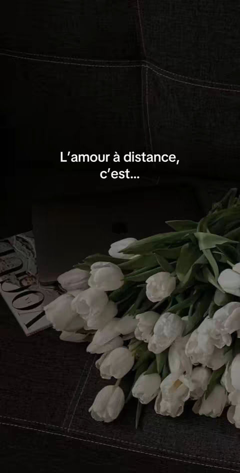 The image showcases a bouquet of white tulips resting on a dark, textured surface, possibly a couch or chair. The composition is simple, with the flowers taking center stage. The background is dark and out of focus, with a magazine visible on the left, creating a soft, moody atmosphere. Text is overlaid, reading "L'amour à distance, c'est..."