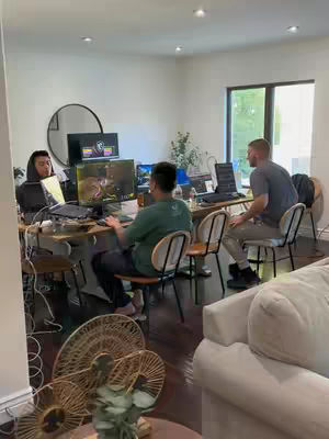 A group of three men working in a modern office setting. Two men are seated at a large desk with multiple computer monitors, while a third man sits to the side. The room has a minimalist design with white walls, dark wood floors, and a window providing natural light.