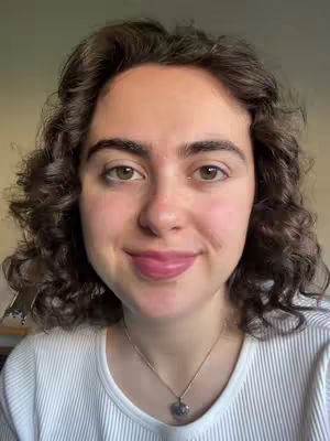 A close-up shot of a young woman with curly brown hair, smiling slightly and looking directly at the viewer. She is wearing a white ribbed shirt and a gold necklace with a small pendant. The background is a blurred, neutral beige.