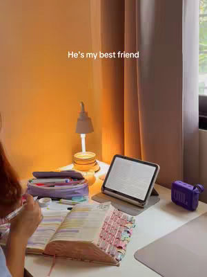 A cozy indoor scene featuring a desk with a Bible, open with highlighted passages and colorful tabs. A pen is poised over the page. Next to the Bible is an iPad, a lamp shedding warm light, and a small speaker. The background includes curtains and a wall. The overall impression is one of study and devotion.