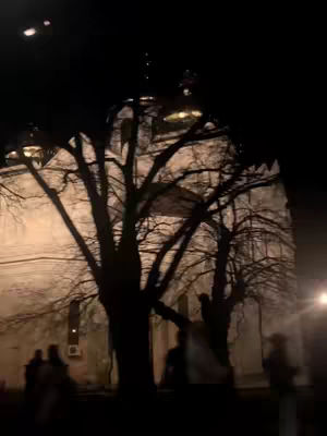 A nighttime shot of an Orthodox church. The church is lit, with its dome visible against the dark sky. In the foreground, bare tree branches create a silhouette. Figures of people can be faintly discerned in the front.