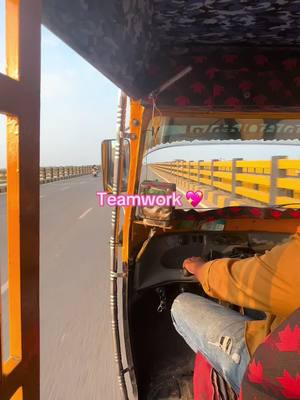 A shot from the inside of a moving vehicle, likely a three-wheeled auto-rickshaw. The view is from the driver's side, showing the steering wheel, part of the driver's body, and the road ahead. A text overlay reads Teamwork with a heart emoji next to it. The background shows a bridge with a yellow barrier on the right side.