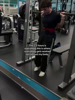 A man is standing in a gym, lifting weights. He is wearing a black t-shirt, black sweatpants with white stripes, and white sneakers. He has a red weightlifting belt around his waist. In the background, there are gym machines and equipment.