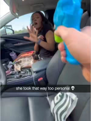A woman is seated in the driver's seat of a car, reacting with shock and wide eyes while holding her hands up. A blue water gun is pointed at her from the right. A text overlay at the bottom reads "she took that way too personal" followed by a skull emoji.