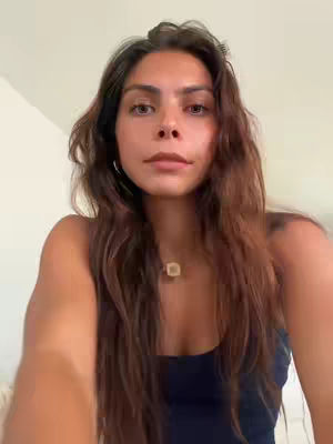 A selfie-style shot featuring a young woman with long, brown hair. She is positioned centrally, gazing directly at the camera with a neutral expression. She wears a dark blue tank top and a gold pendant necklace. The background is a plain, light-colored wall, which directs focus to the woman. The lighting is soft and diffused, without harsh shadows.