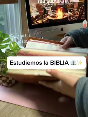 An image showing a person holding an open bible, with the text overlay saying, 'Estudiemos la BIBLIA'. The background shows a computer screen displaying an image of Adirondack chairs near a fire, and a window with sunlight.