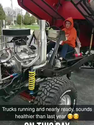 A person sits in the partially assembled chassis of a vehicle, likely a truck. The color scheme is dominated by blacks, silvers, and reds of the vehicle components. The person, dressed in an orange hoodie, looks towards the camera. The background shows trees and greenery, suggesting an outdoor setting, possibly a yard or driveway.