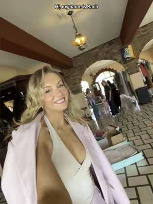 A selfie-style video shot in what appears to be a large indoor space, possibly a home or studio. The primary subject is a woman with blonde hair, wearing a light-colored outfit and a pink blazer. She is smiling at the camera. The background is somewhat blurred, showing architectural details like brickwork and arches, with a few people visible in the distance. The overall composition is well-lit, with a focus on the woman's face and expression, suggesting a casual, personal tone. There is text at the top of the image stating 'Hi, my name is Rach'.