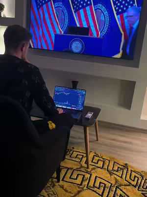 A man is seated in a living room, facing away from the camera, focused on a laptop displaying a financial chart. A large TV screen in the background shows a room with American flags, suggesting a government setting. The room has contemporary decor with a patterned yellow rug.