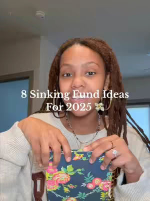 A woman with brown locs is centered in the frame, holding a floral notebook. Text overlays on the image read "8 Sinking Fund Ideas For 2025". She wears a cream-colored sweater, a necklace, and rings. The background is slightly blurred.