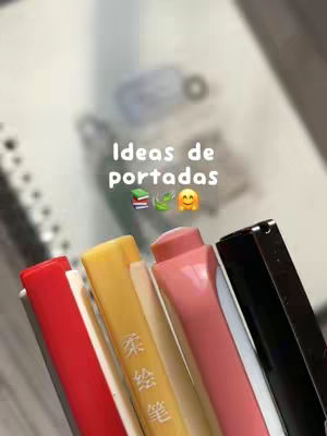 The image shows a close-up of several colorful pens and markers, with a blurred background that suggests notebooks. In the center, the text "Ideas de portadas" is displayed in white with associated emojis. The pens are arranged diagonally across the bottom, with red, white, yellow, pink, and black visible. The layout is clean, focusing on the stationery and the text overlay.