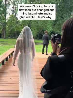 A woman in a wedding dress walks away from the camera on a wooden deck towards a man in a suit. A person with a camera is partially visible on the right side of the frame. Text overlay at the top says: "We weren't going to do a first look but changed our mind last minute and so glad we did. Here's why..."