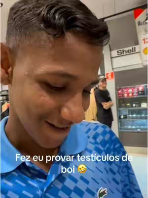 A young man is looking down and slightly to the left, mid-smile, with a blue polo shirt that has a repeating pattern of an island pattern. He is in what looks like a store with a Shell sign in the background.