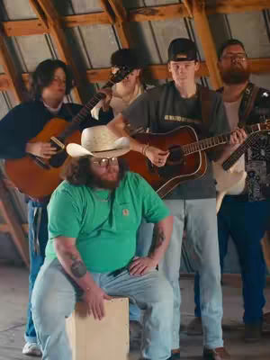 A group of musicians is posing for a photo in what looks like an attic setting. The lighting is warm, with natural light coming from the side. The color palette is muted, warm and natural. The composition is a medium shot, showing all the musicians.