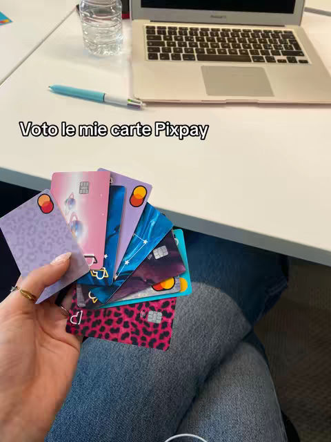 A person's hand holds a fan of colorful credit cards in front of their lap, with a laptop and other items on a white surface visible in the background. The hand is in the lower left holding a varying array of cards, from blue to pink to purple with different designs, while the right side of the image shows part of a laptop, a pen, and a bottle of water.