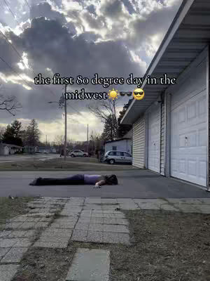 A woman lays face down on a dark asphalt road. Around her is concrete and a garage. The sky is dramatic.