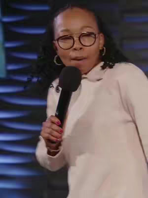 A medium shot shows a woman holding a microphone, facing slightly off-center. She is wearing round glasses, hoop earrings, and a light-colored sweater. Her expression suggests she is speaking or singing. The background is a textured blue surface.