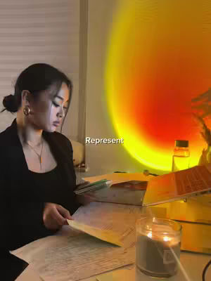 A young woman with dark hair pulled back into a bun sits at a desk, papers and a candle present.  A soft, warm light, mimicking a sunset. She is focused on reading.