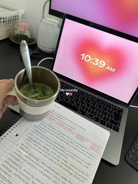 A well-lit workspace featuring a laptop displaying a blurred heart-shaped image with the time "10:39 AM" on its screen. A mug, holding a green beverage, is in the foreground. Handwriting with organized notes is visible on a spiral notebook, and the scene conveys a study or work-from-home environment.