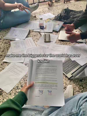 The image shows a group of people studying, with papers and a laptop scattered around them. One person holds an open book, and there's a bottle of water and a phone on the ground.