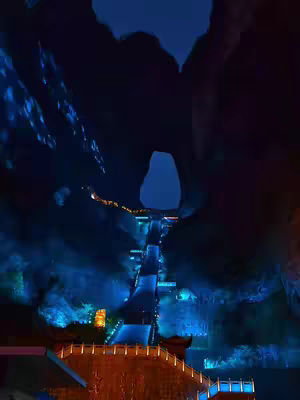 A nighttime shot of what appears to be a scenic location with a road or pathway winding upwards beneath a dark, arched opening. The scene is illuminated by blue and orange lights, creating a dramatic and somewhat mystical atmosphere. The composition leads the eye upwards, with a structural element in the foreground.