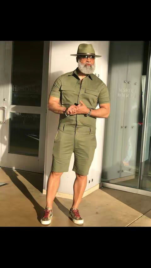 A man with a gray beard and mustache poses outdoors in a green jumpsuit, hat, and sneakers. The background includes a building facade and a glass door.