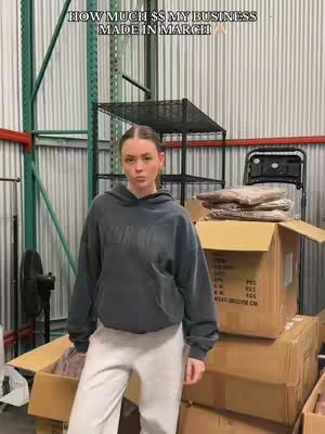 A woman wearing a hoodie and light pants stands in front of boxes and storage units. Text at the top reads, "HOW MUCH $$ MY BUSINESS MADE IN MARCH yayyy"