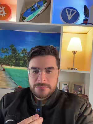 A man with glasses and a mustache is holding a microphone. He is in front of a shelf filled with objects, including a blue painting, a surfboard, and a sign with a bitcoin symbol.